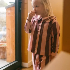 Child wearing TBCo Cotton Kids Pyjamas in Pink & Burgundy Stripe standing by glass door in a cosy indoor setting
